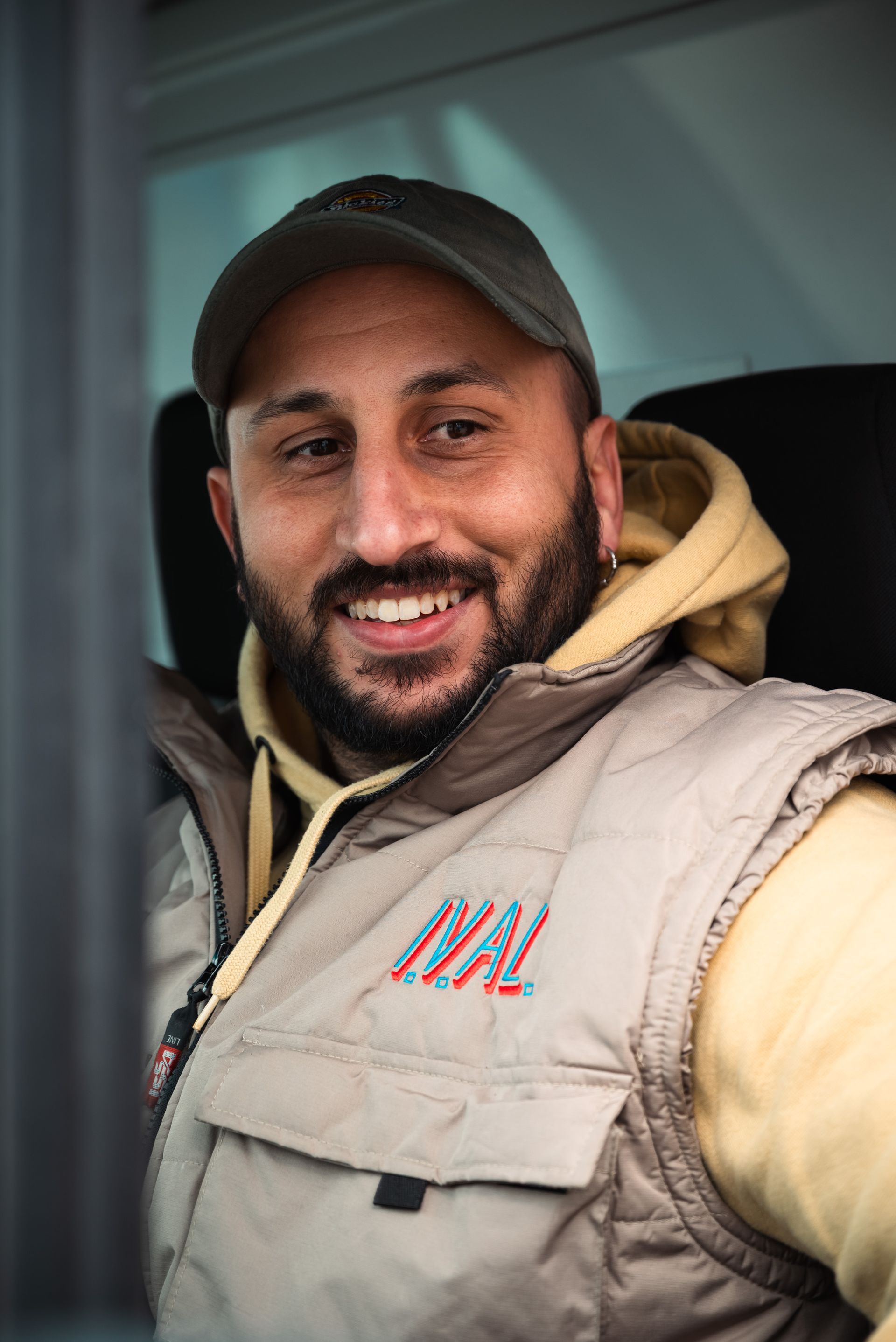 Un individuo sorridente, con indosso un gilet beige sopra una felpa con cappuccio di colore chiaro e un berretto da baseball verde, si trova all'interno di un veicolo.