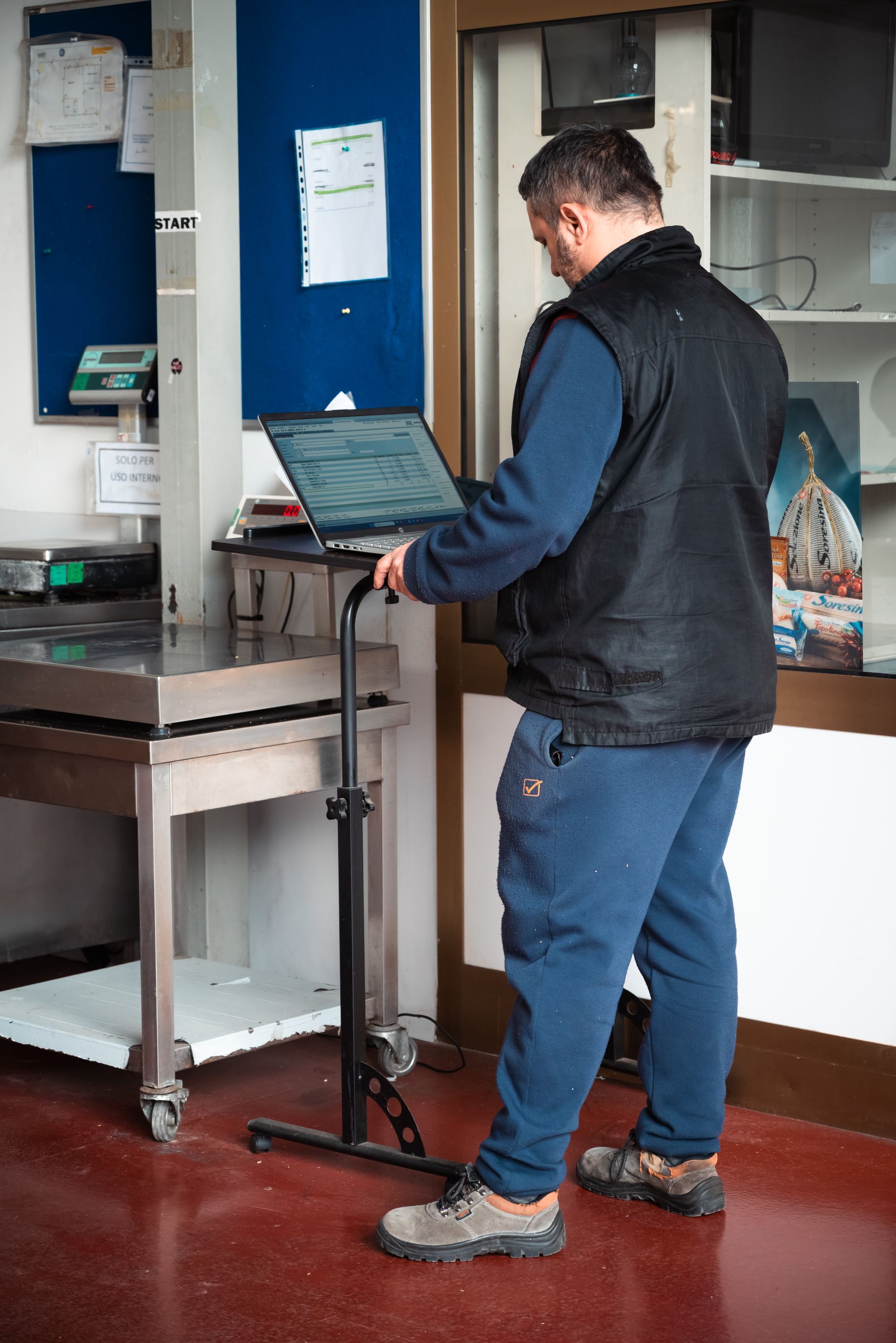Una persona con un gilet nero e pantaloni della tuta blu è in piedi accanto a un alto supporto portatile per laptop in una postazione di lavoro vicino a una bilancia metallica.