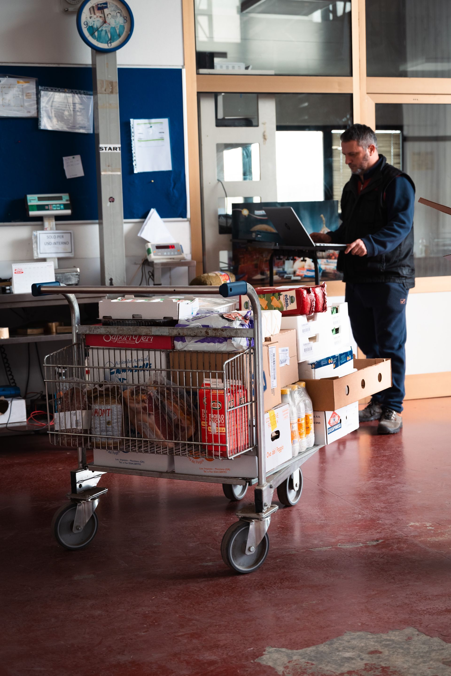 Un operaio si trova in un locale commerciale, intento a usare un computer portatile mentre è appoggiato a un grande carrello metallico carico di merci imballate.