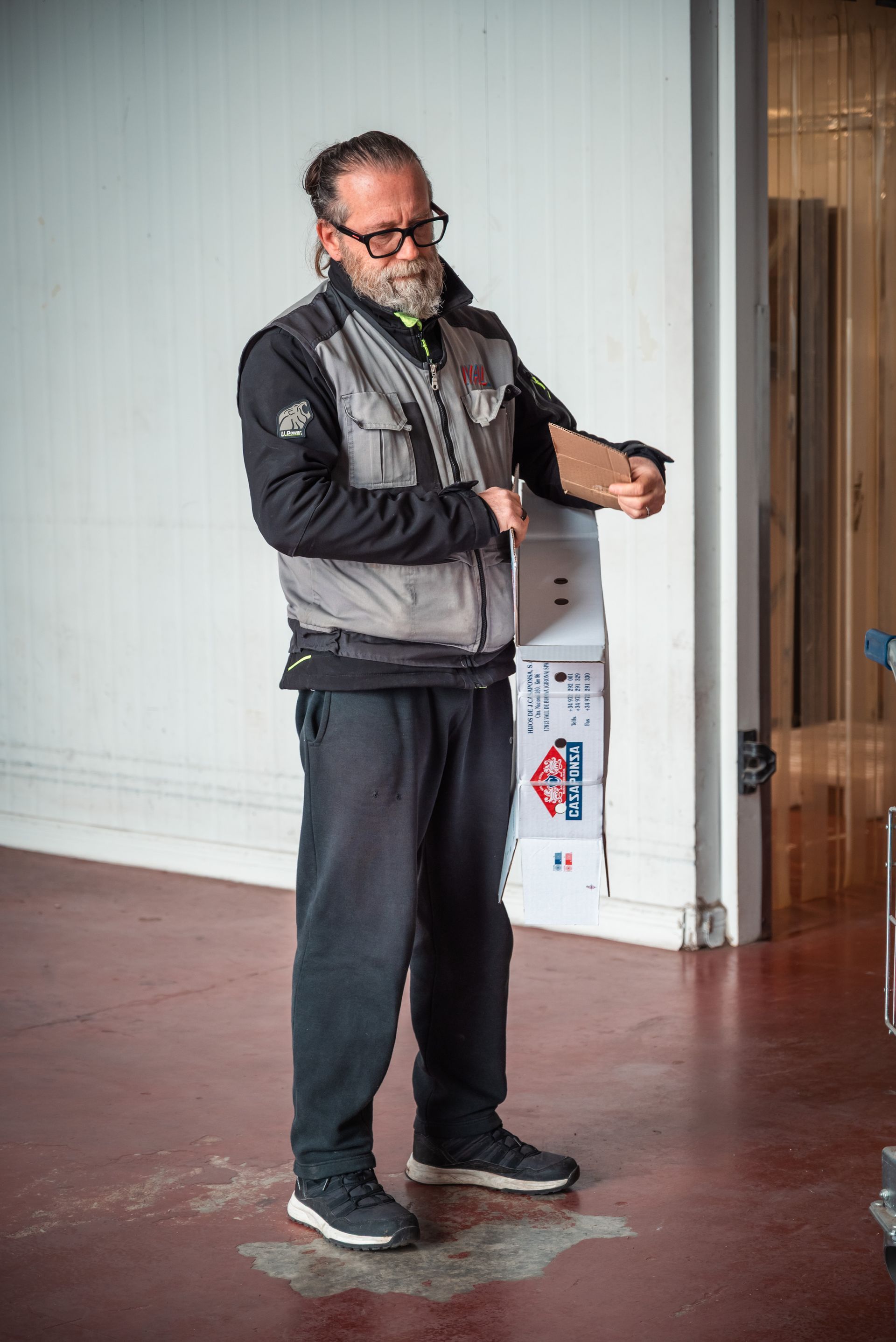 Un uomo barbuto con una giacca da lavoro grigia e pantaloni scuri è in piedi su un pavimento rossastro, con in mano una scatola di cartone.