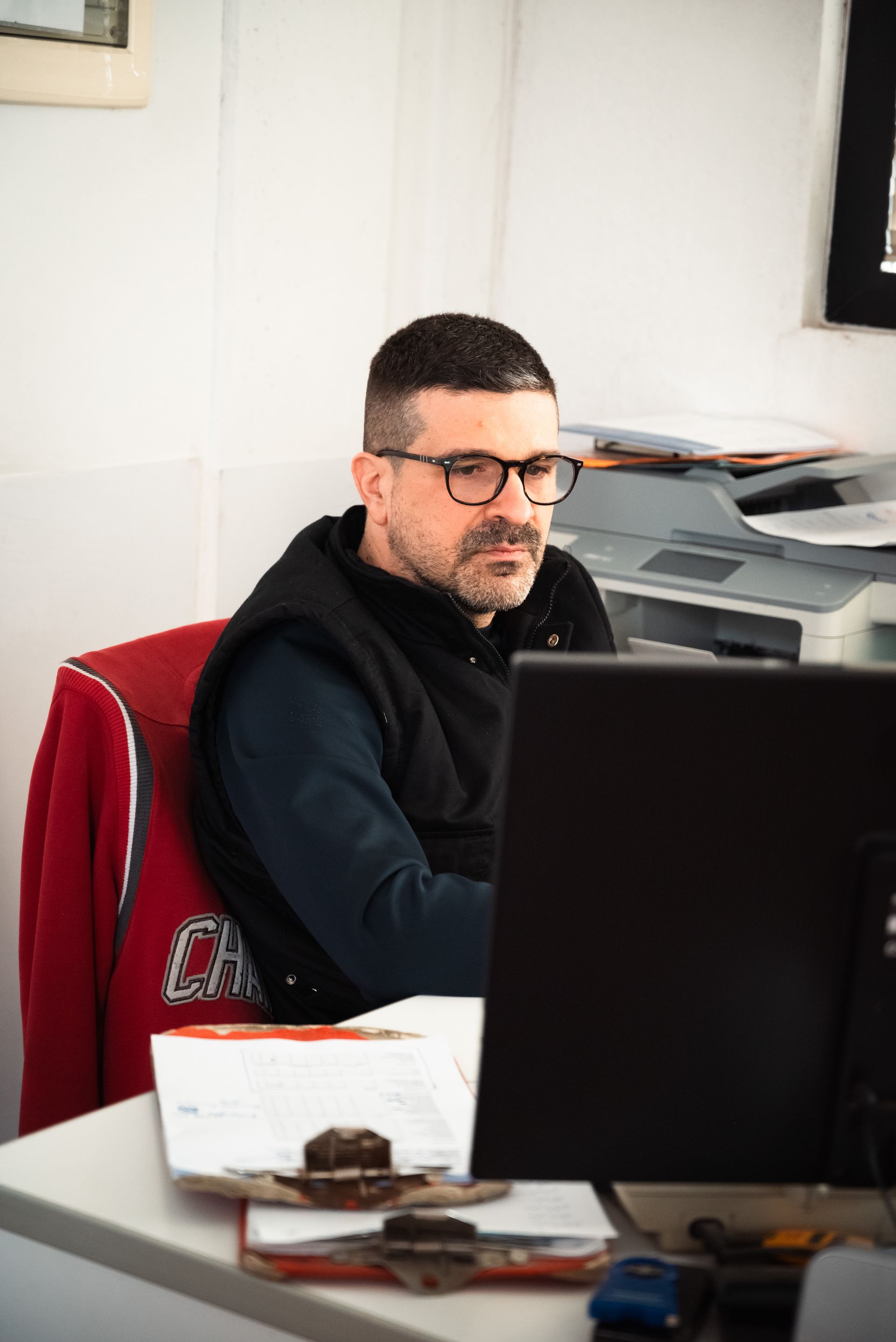 In un ufficio luminoso, una persona con gli occhiali e un gilet nero lavora a una scrivania con un computer, una stampante e dei documenti.
