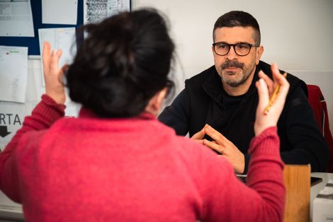 Durante una riunione in ufficio, una persona con un maglione rosso gesticola mentre parla con un uomo che indossa un maglione scuro e gli occhiali.