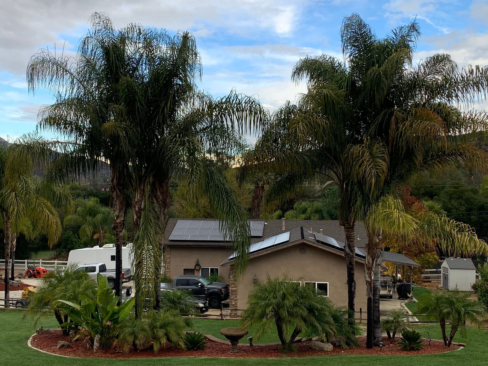 House with palm trees, green lawn, and vehicles, under a cloudy sky.