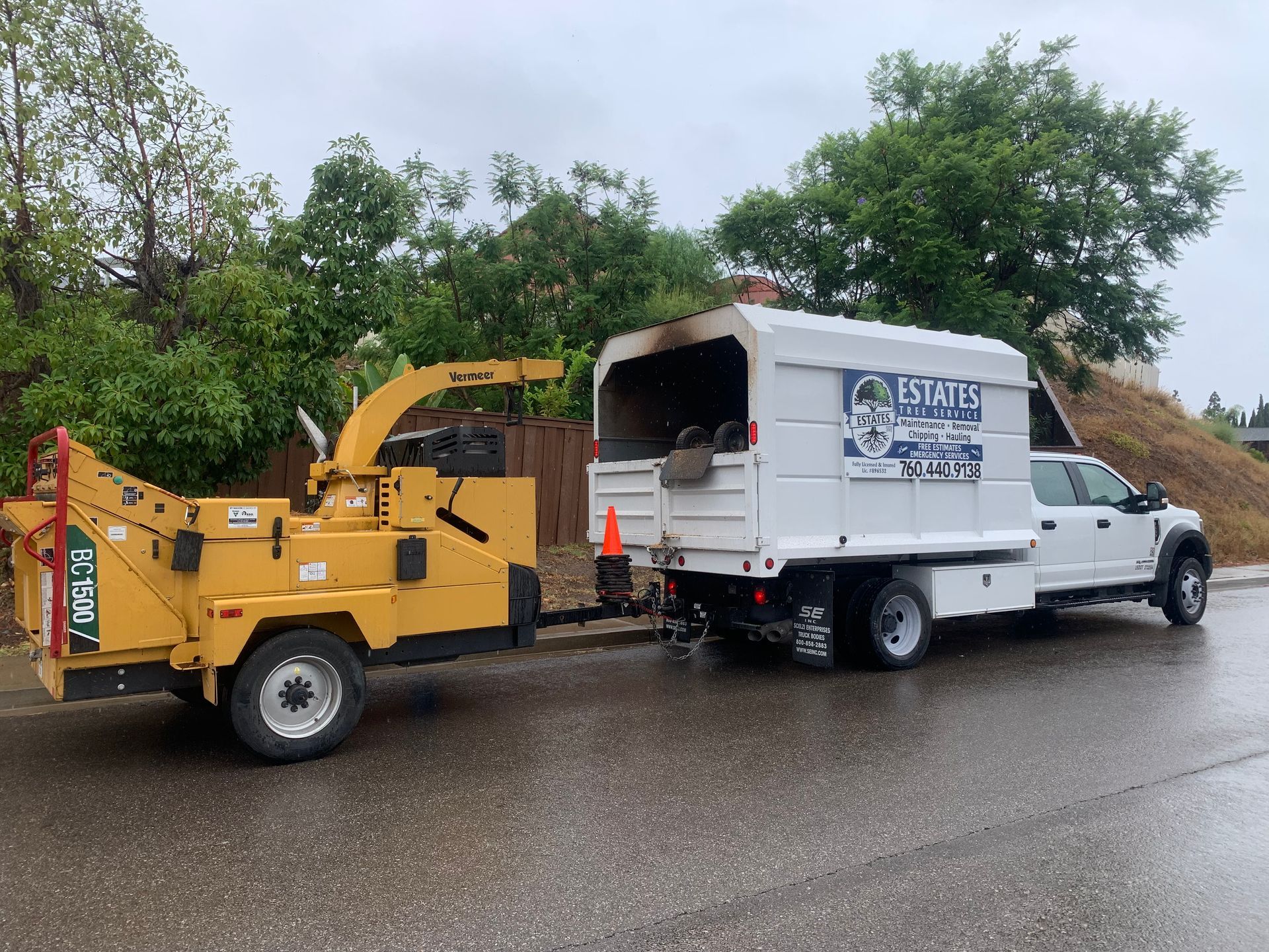 Yellow wood chipper trailer hitched to a white truck on a wet road; green foliage in background.