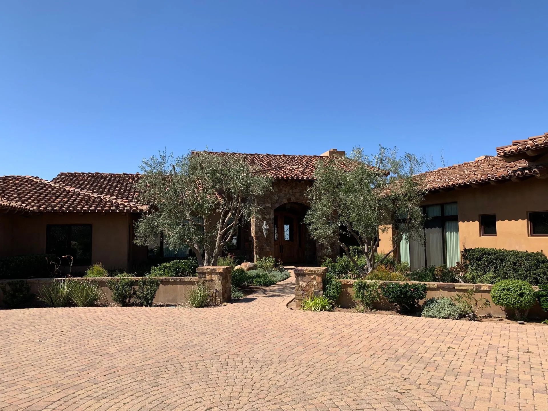 A single-story, tan stucco house with a tiled roof and circular brick driveway under a clear blue sky.
