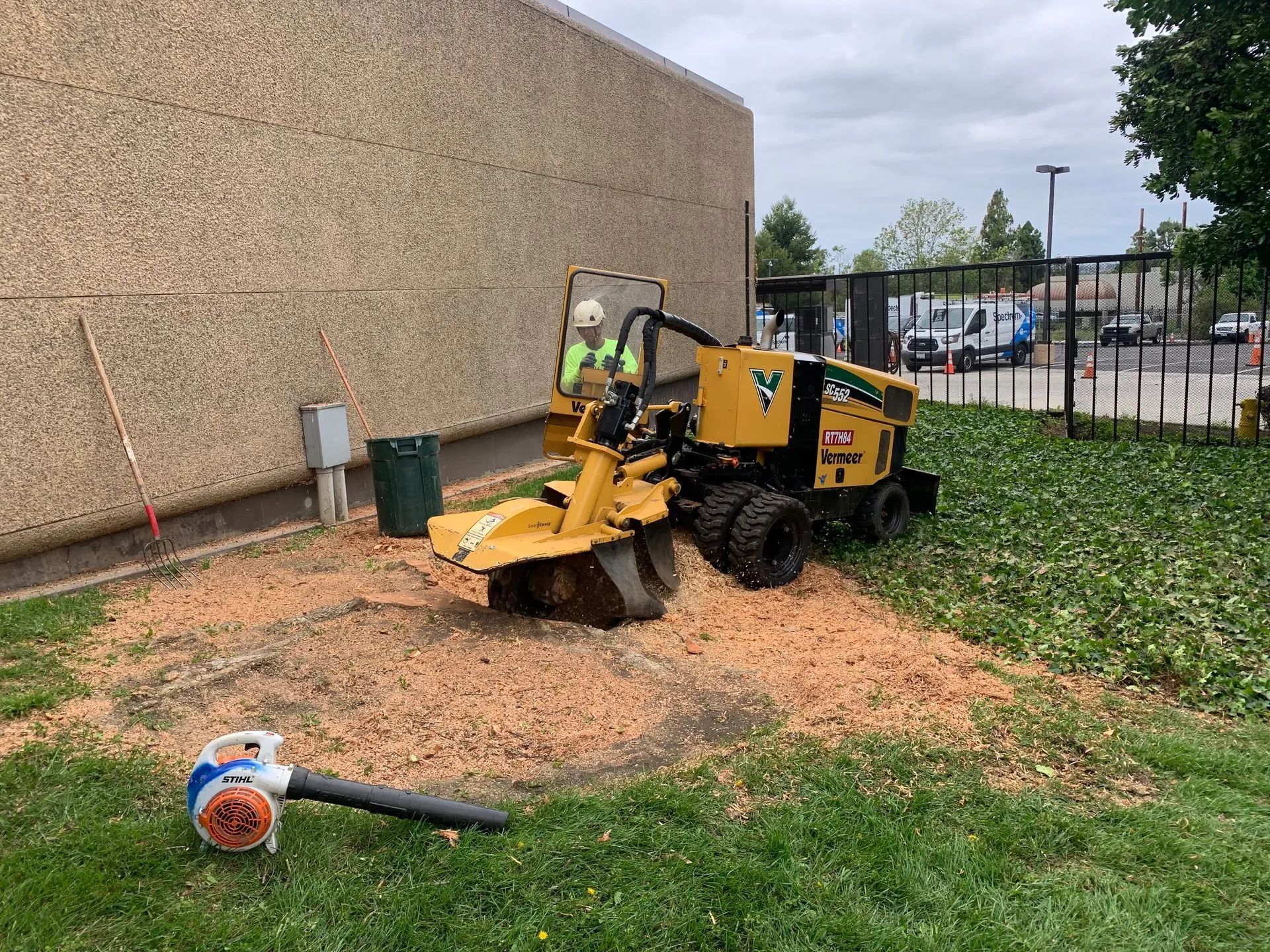 Man operating a stump grinder on a lawn near a building. Brown wood chips scattered around.