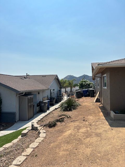 Buildings with brown dirt path, waste bins, and landscaping. Mountain in the background under a blue sky.