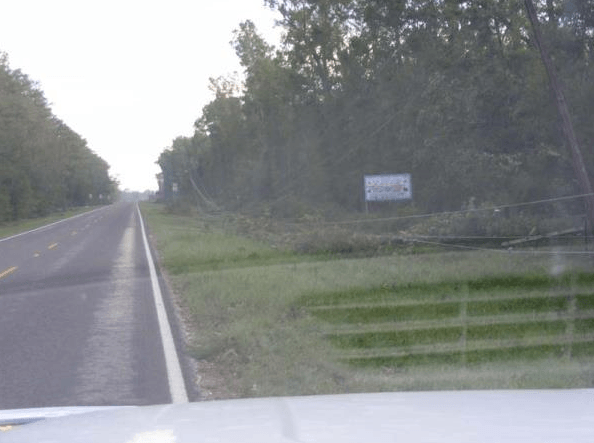 Straight rural road with trees on both sides and a roadside sign in the distance.