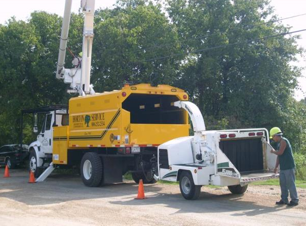 Yellow utility truck hoisting a white pickup beside a worker and traffic cones on a roadside