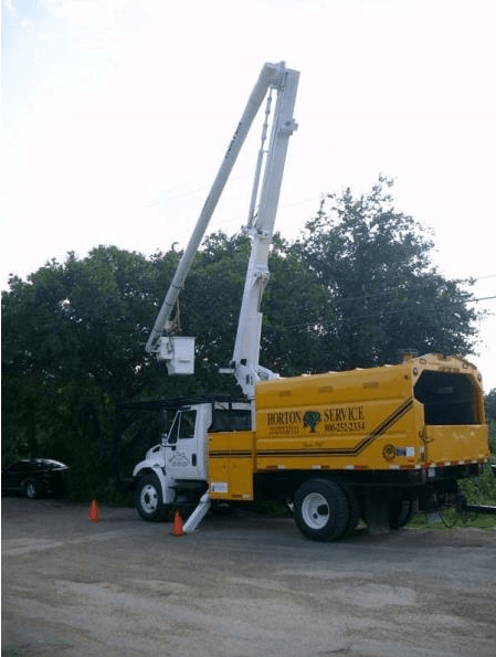 Yellow utility bucket truck with raised boom parked beside trees on a road, cones nearby