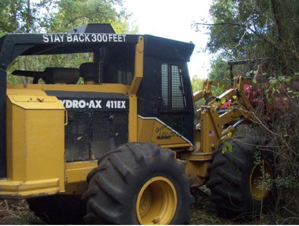 Yellow Hydro-AX 4x4 tractor pushing through brush in a wooded area, with “STAY BACK 300 FEET” sign.