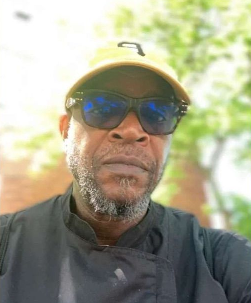 Man wearing sunglasses and a yellow cap, with a graying beard, in a chef's uniform.