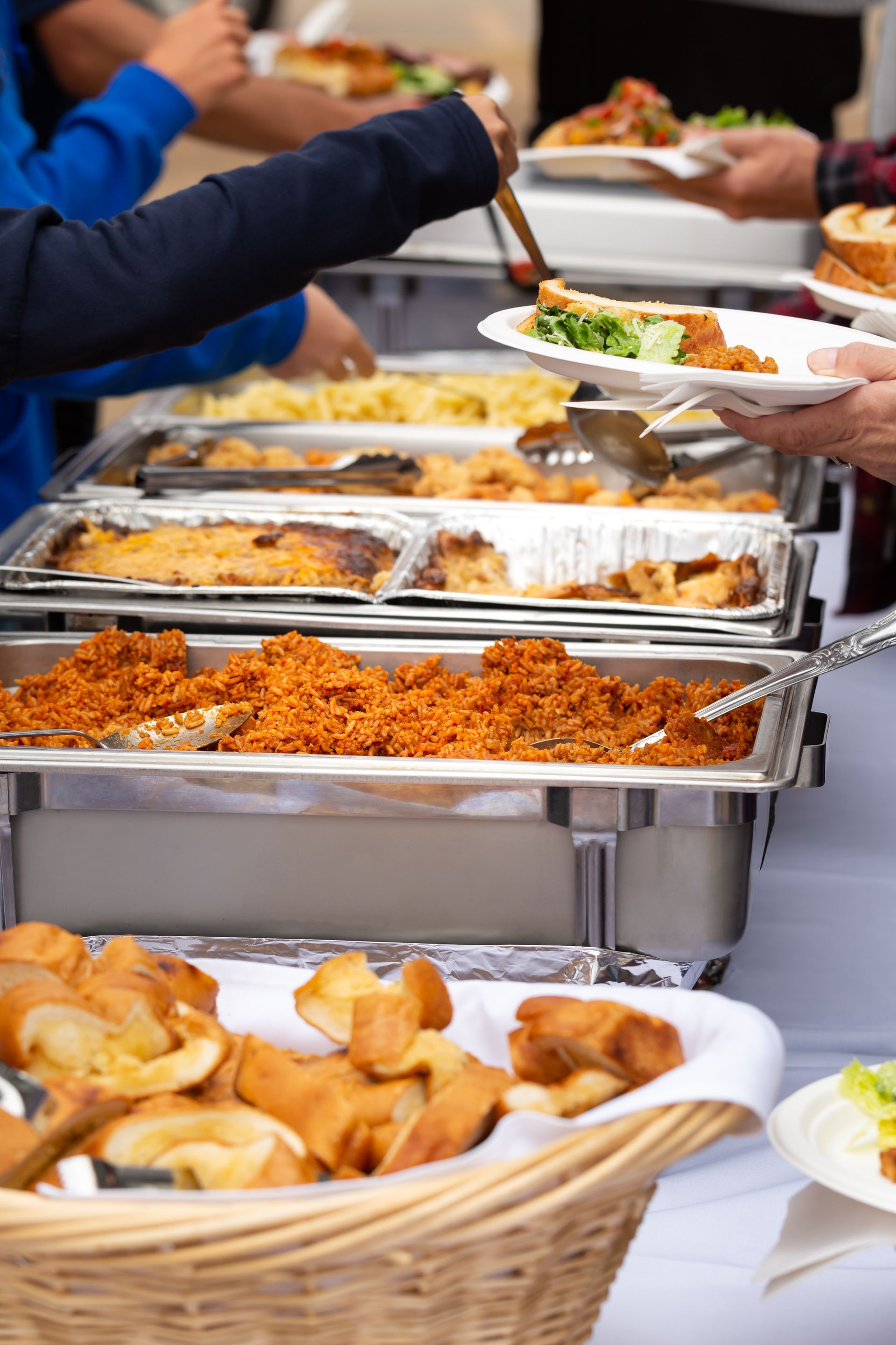 People serving themselves from a buffet table with various dishes like pasta, rice, and bread rolls.
