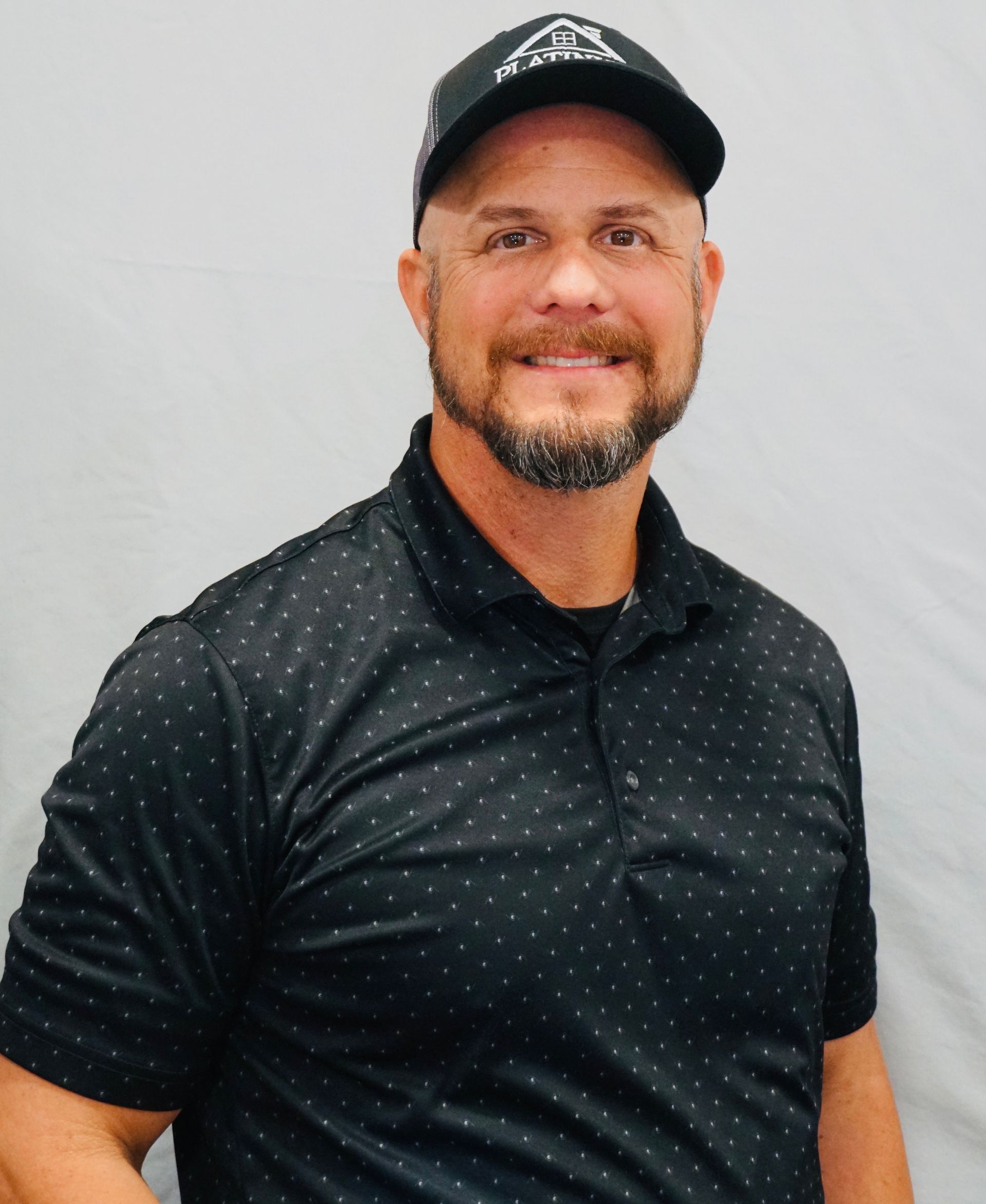 A man with a beard is wearing a black polo shirt and a black hat.