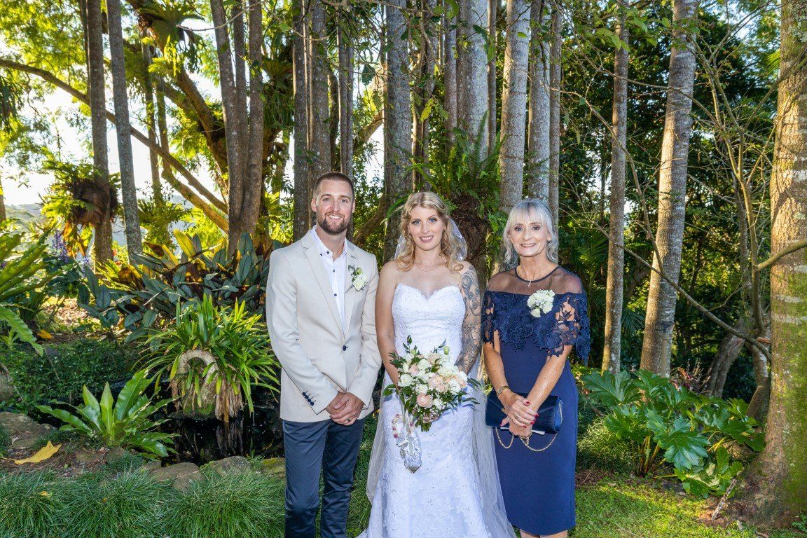 a bride and groom are posing for a picture with their mother in front of a forest .