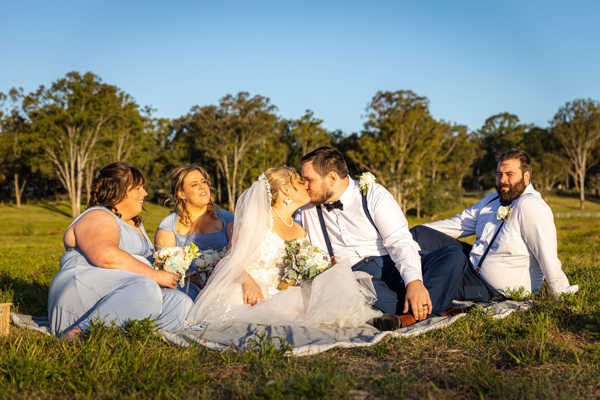 a bride and groom are kissing while sitting on a blanket with their wedding party .