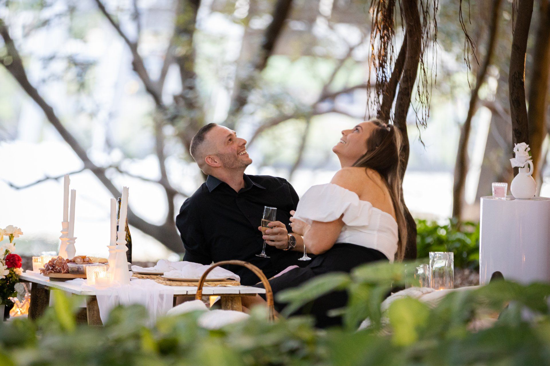 a man and a woman are sitting at a table under a tree holding champagne glasses .