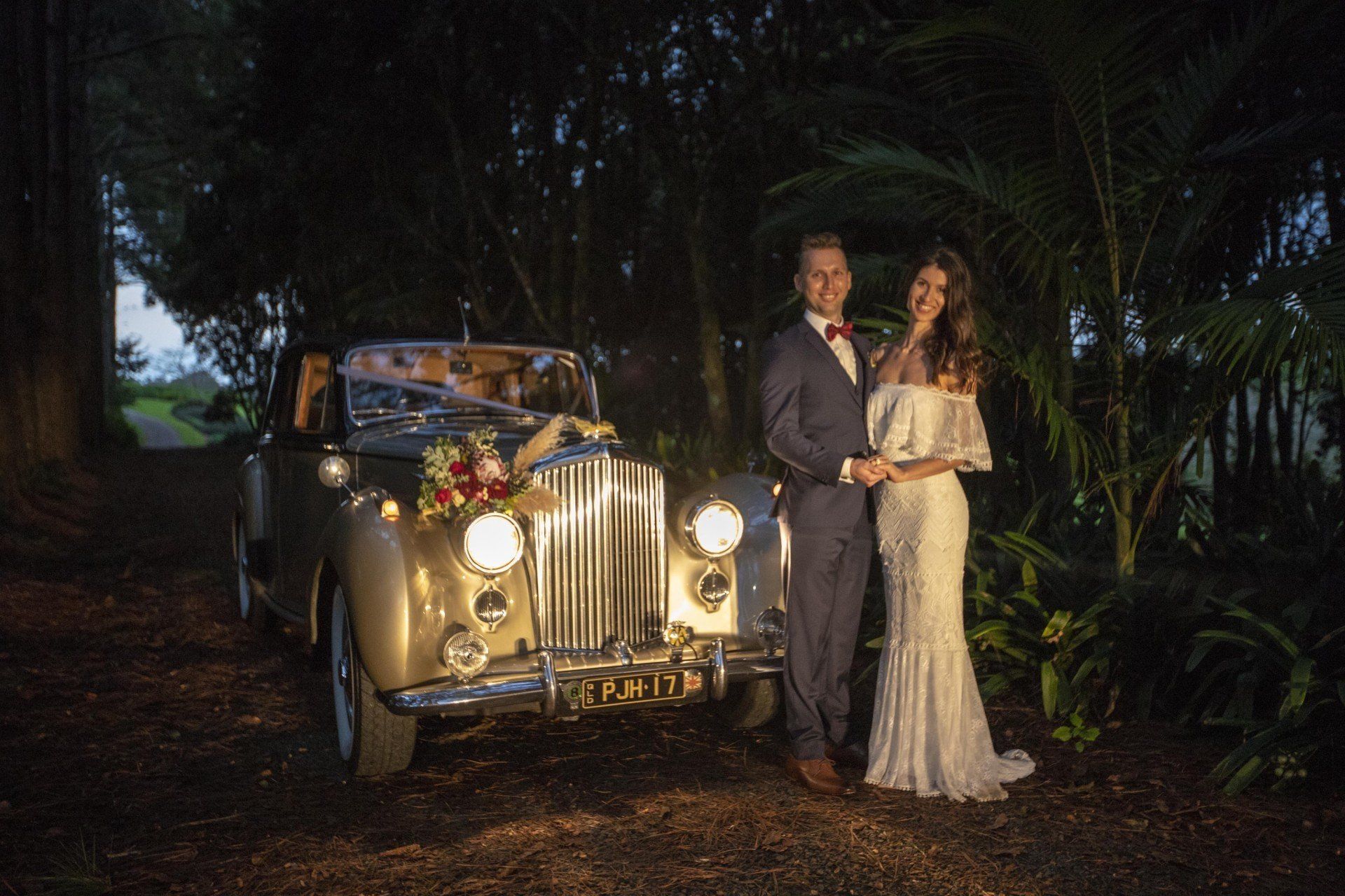 a bride and groom are posing for a picture in front of a vintage car .
