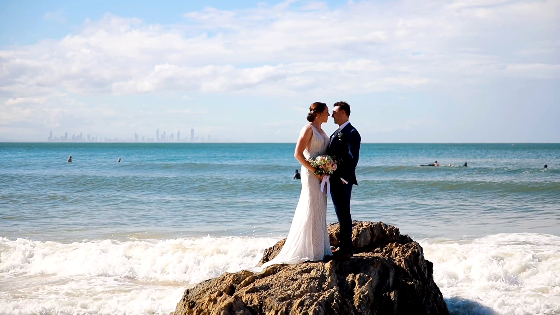 a bride and groom are standing on a rock near the ocean .