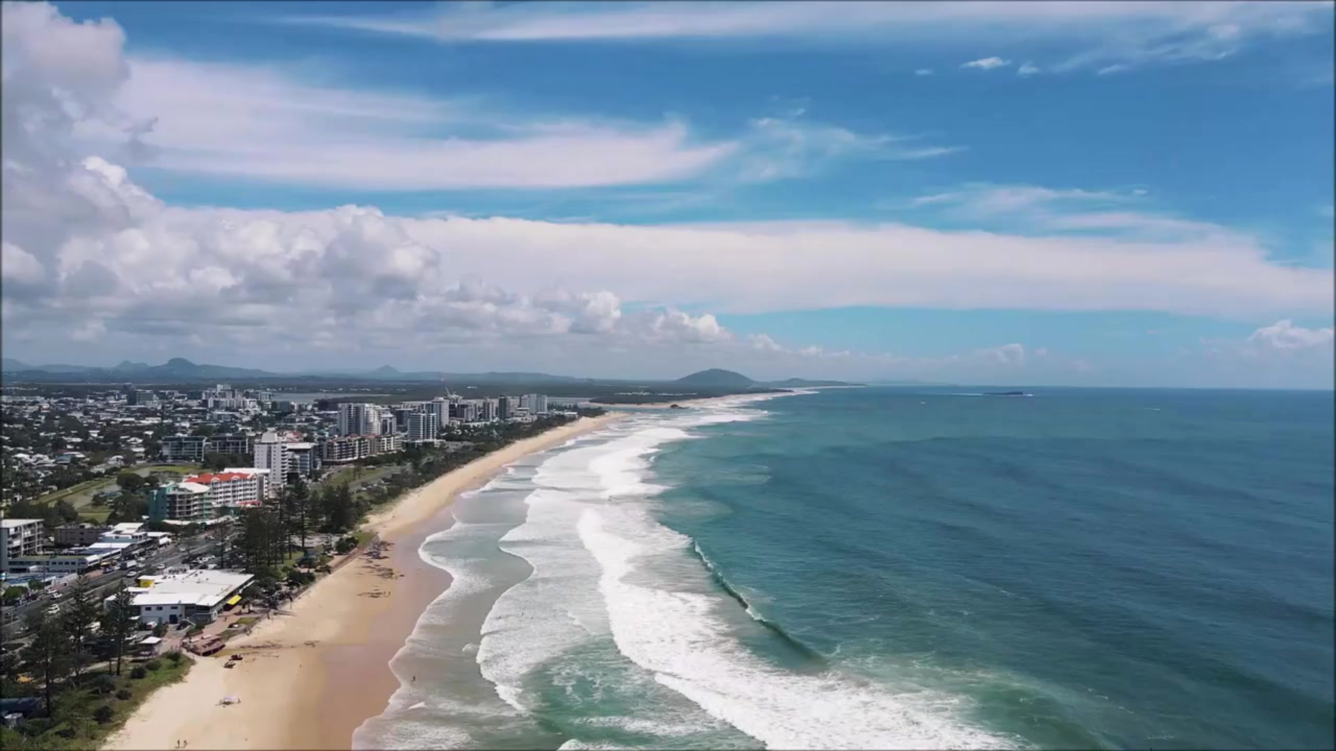An aerial view of a beach with waves and a city in the background.