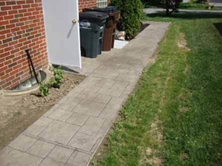 A sidewalk leading to a brick building with trash cans on the side of it.