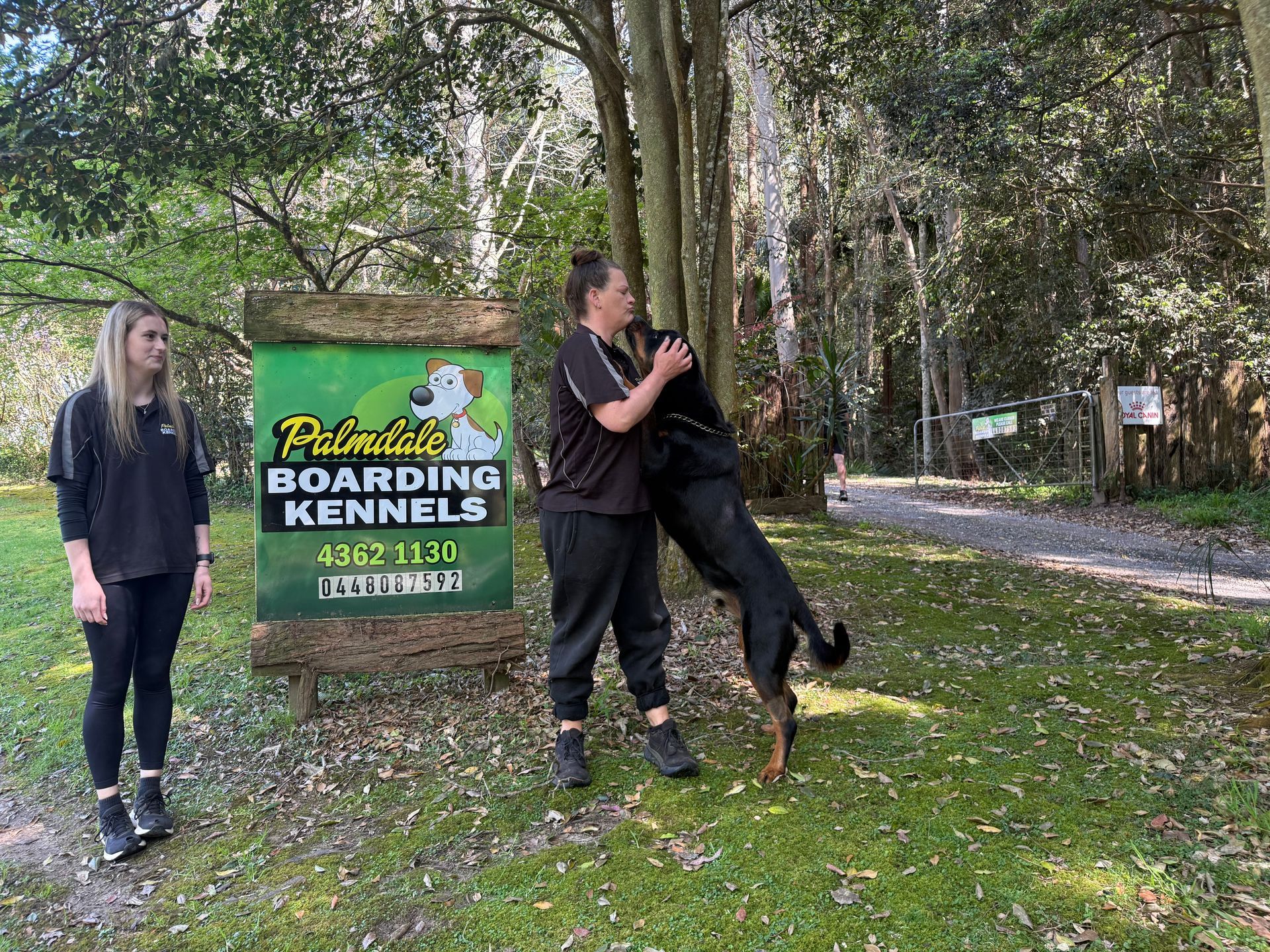 Two people and a dog at Palmdale Boarding Kennels sign. Woman hugs dog, other woman looks on. — Palmdale Boarding Kennels In Palmdale, NSW