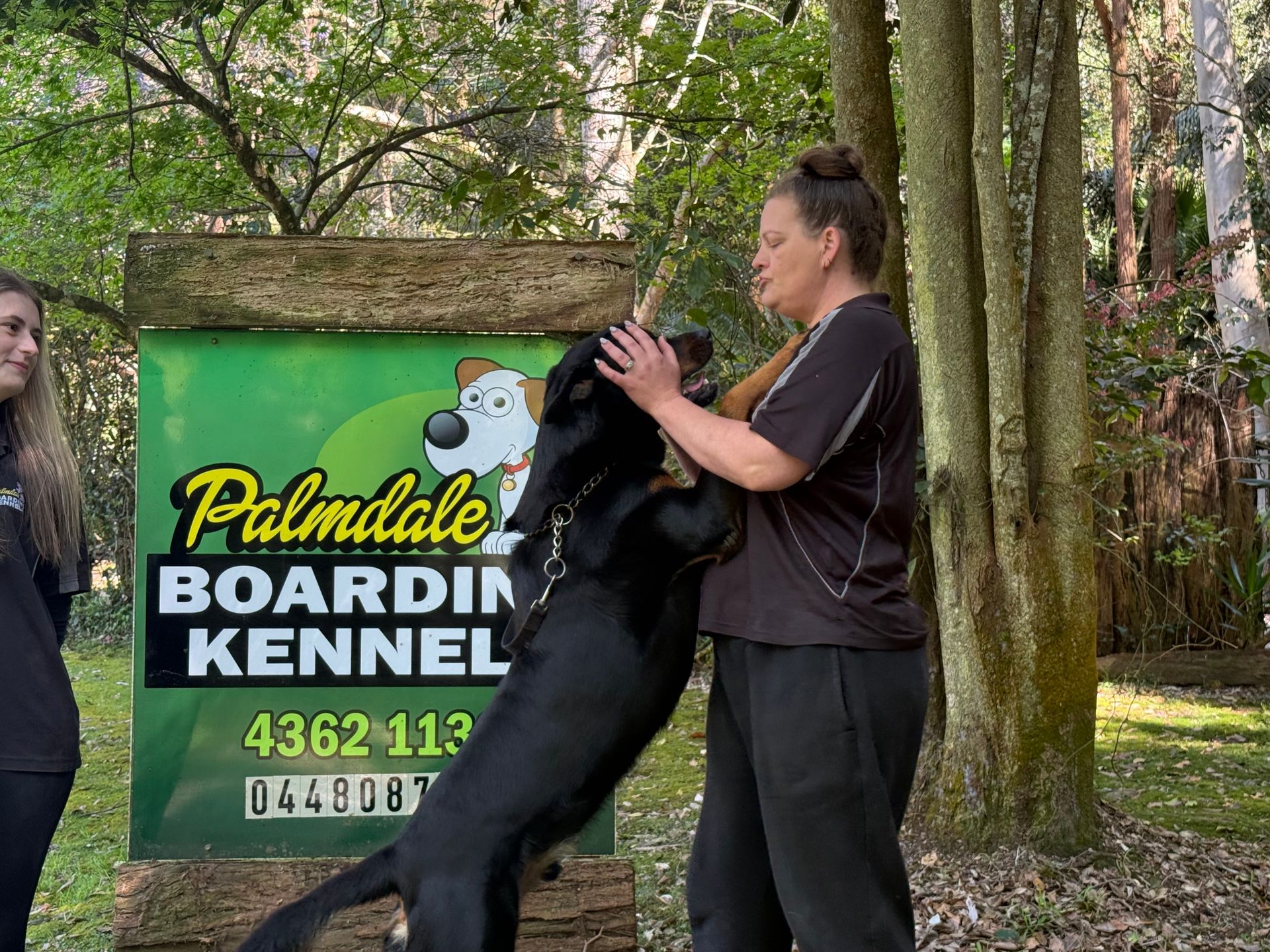 Woman petting a black dog in front of a Palmdale Boarding Kennel sign; other person watches.— Palmdale Boarding Kennels In Palmdale, NSW