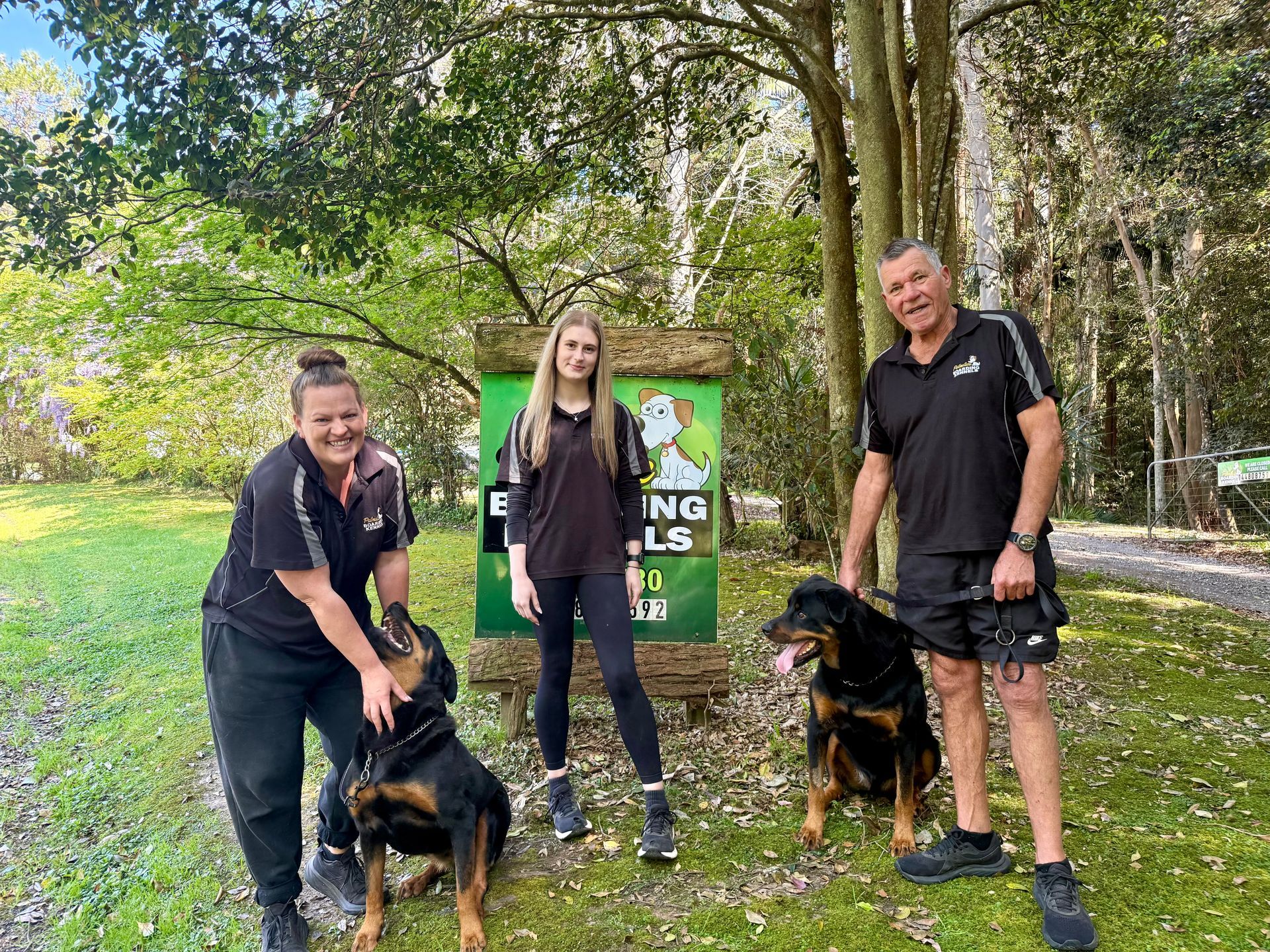 Three people and two Rottweilers pose in front of a sign in a park.— Palmdale Boarding Kennels In Palmdale, NSW