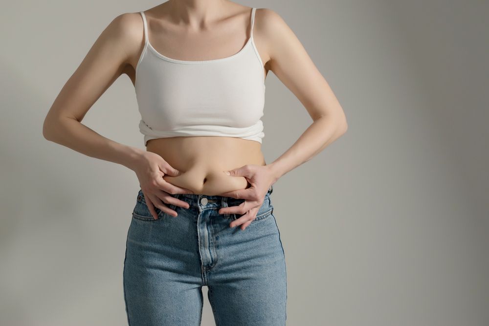 A woman in a white tank top and blue jeans is holding her stomach.