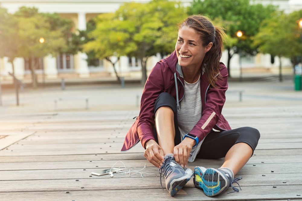 Woman in athletic wear smiles while tying shoelaces on a wooden surface, outdoors in a city square.