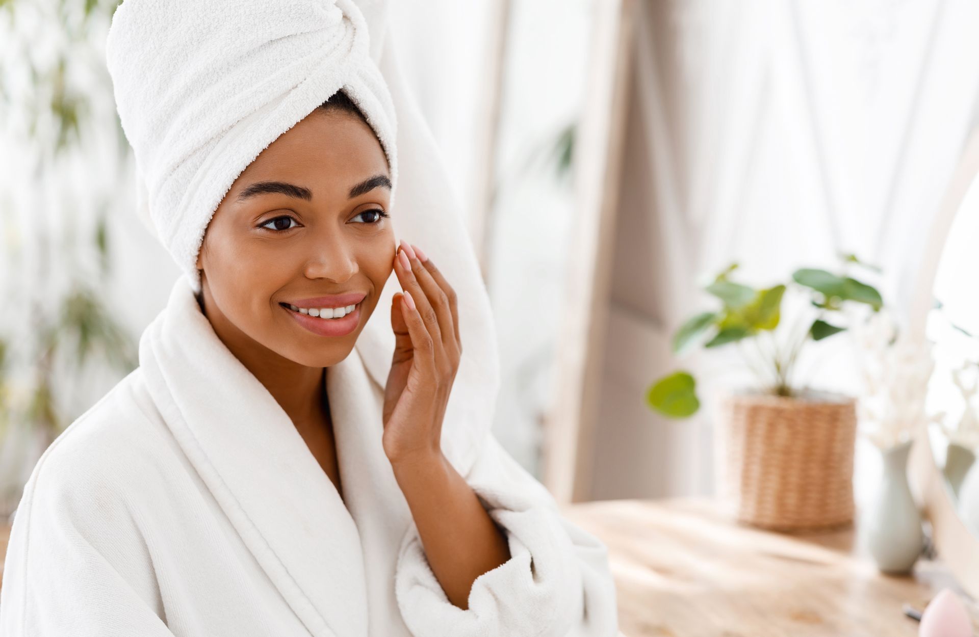 A woman with a towel wrapped around her head is touching her face in front of a mirror.