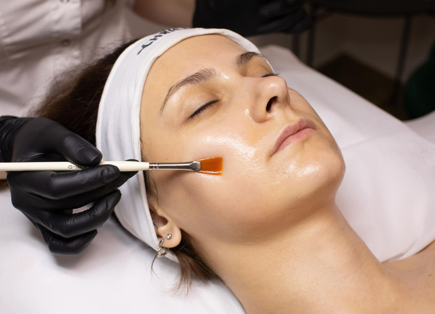 Woman receiving a facial peel treatment. A professional applies liquid with a brush to her cheek, in a salon.
