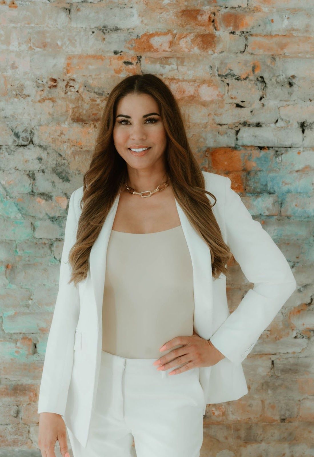 A woman in a white suit is standing in front of a brick wall.