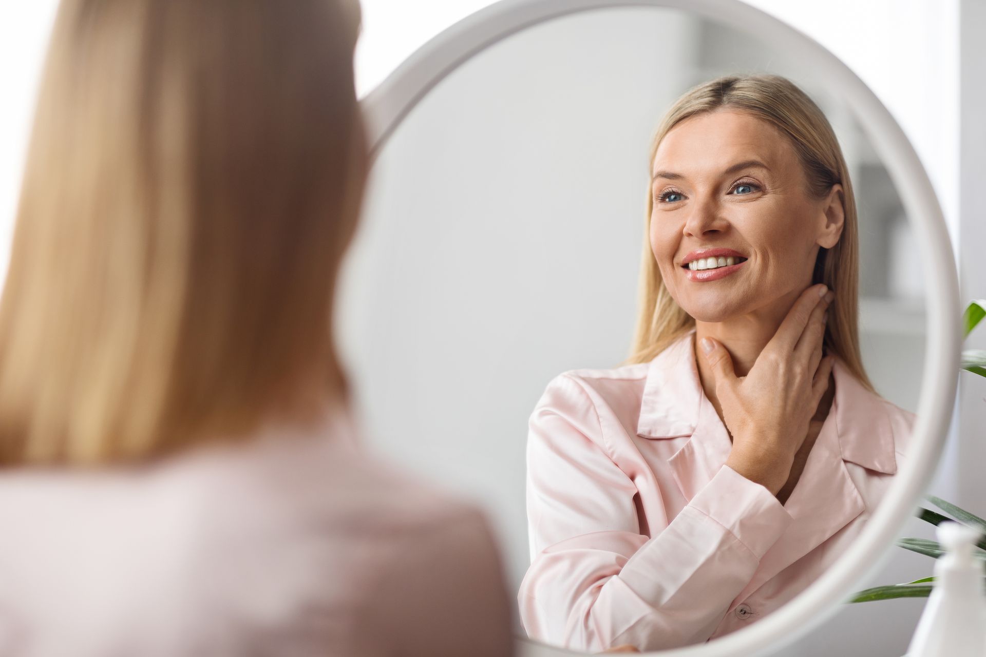 Woman looking in mirror, touching her neck, smiling. Pink shirt, round mirror in bathroom.