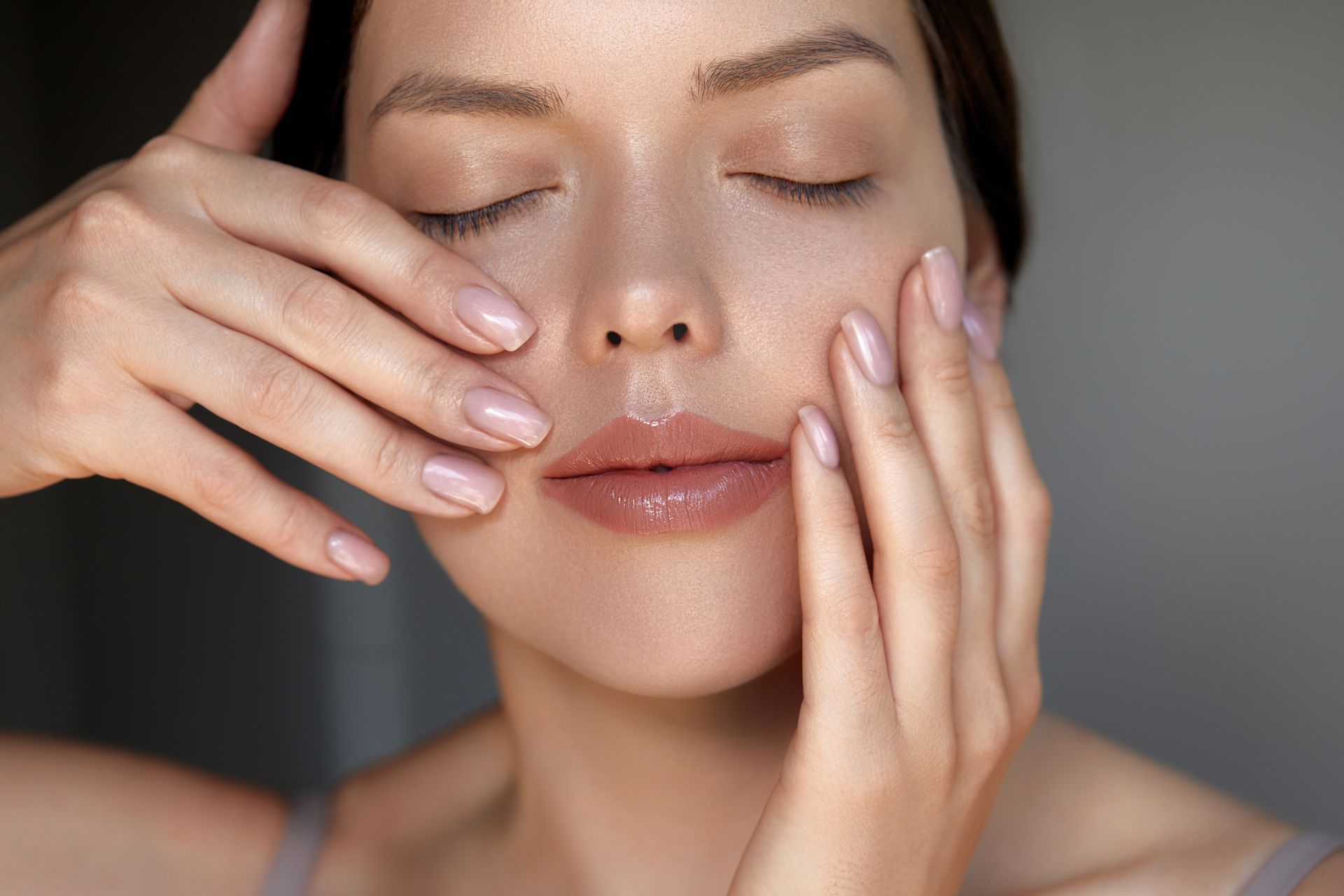 Woman with eyes closed, hands on face, appears to be applying product, soft lighting.