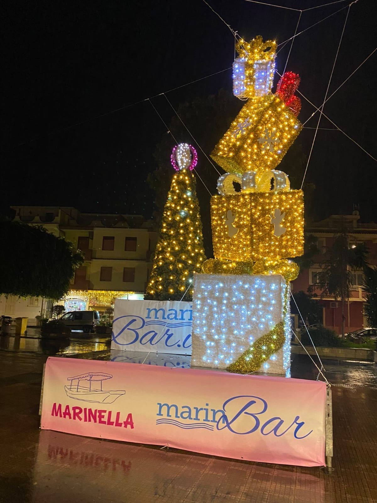 Sculture decorative illuminate e scatole regalo in una piazza cittadina di notte, con l'insegna del Marinella Bar.