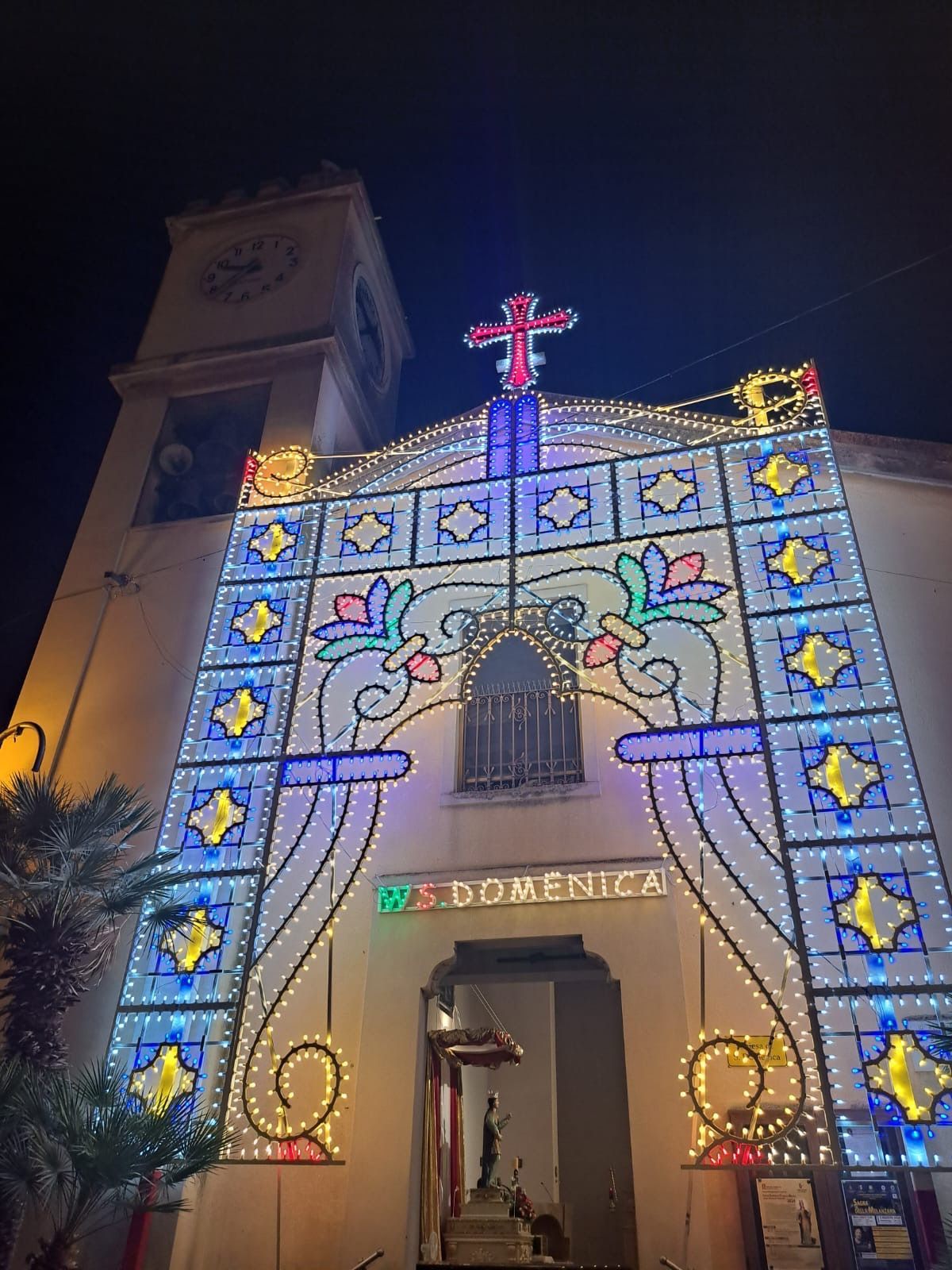 L'ingresso della chiesa illuminato di notte con luci colorate a forma di stelle e fiori, il portale ad arco e la croce in cima alla facciata
