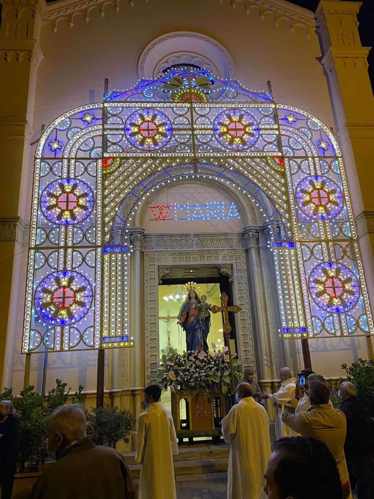Santuario illuminato con luci blu e rosa, una statua centrale di un santo e persone riunite sotto di notte.