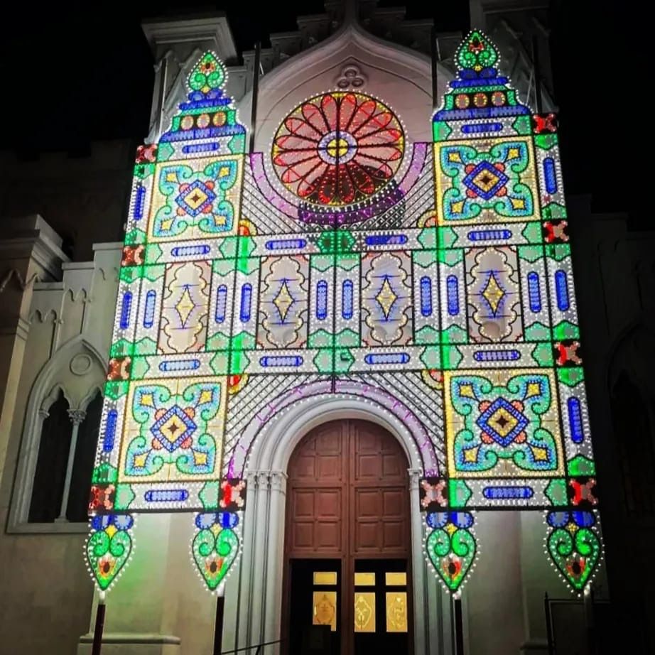 Di notte, una facciata colorata e illuminata con motivi a vetrata artistica attorno a un portale ad arco in legno.