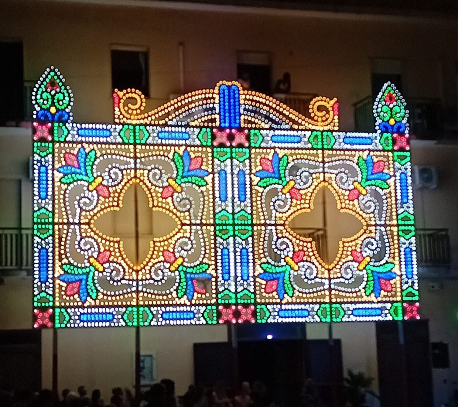 Di notte, un edificio è illuminato da un'esplosione di luci colorate in stile vetrata, con motivi floreali e geometrici simmetrici.