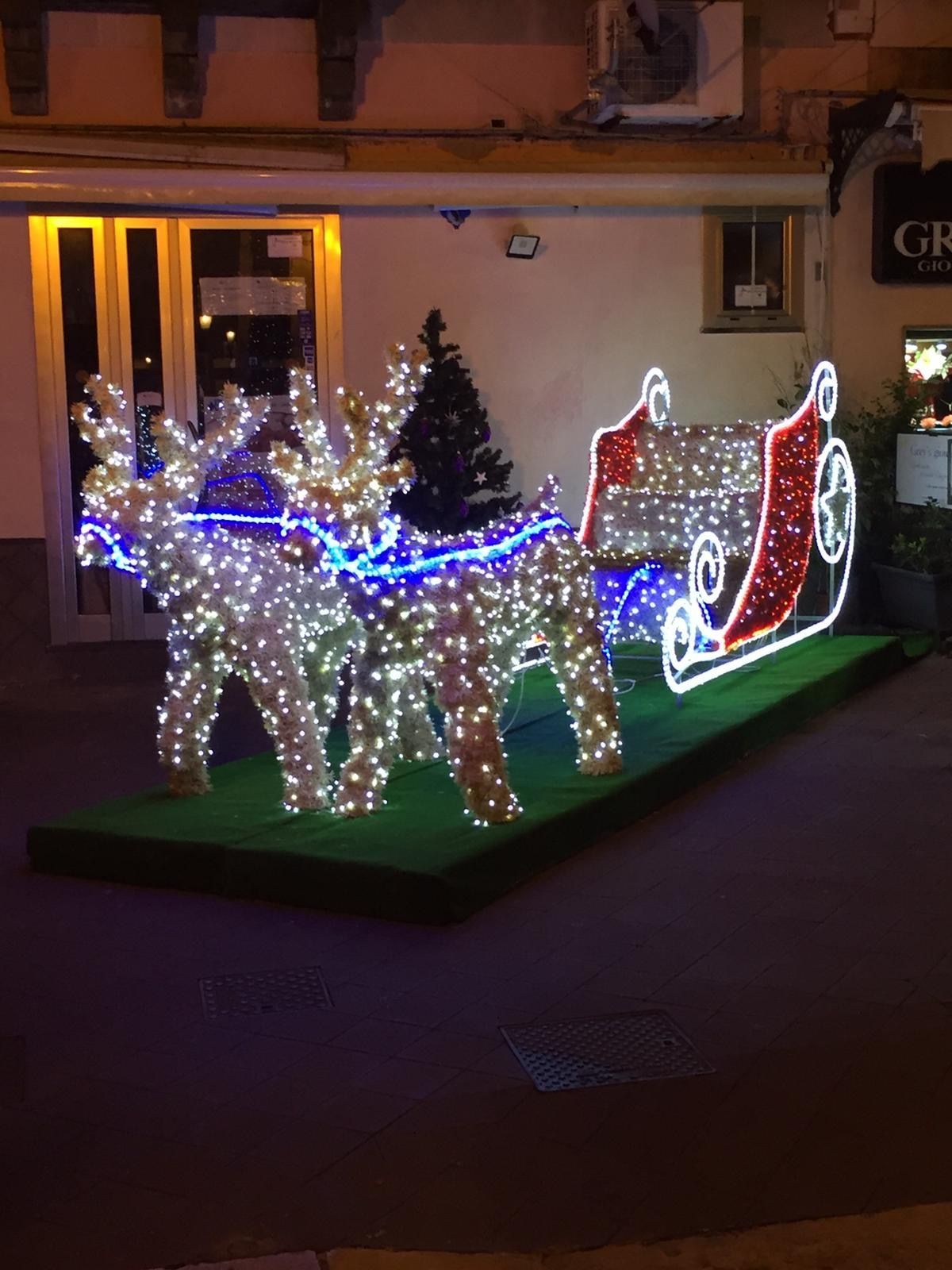 Decorazione natalizia illuminata con renne e slitta, con luci blu, bianche e rosse, all'esterno di un edificio.