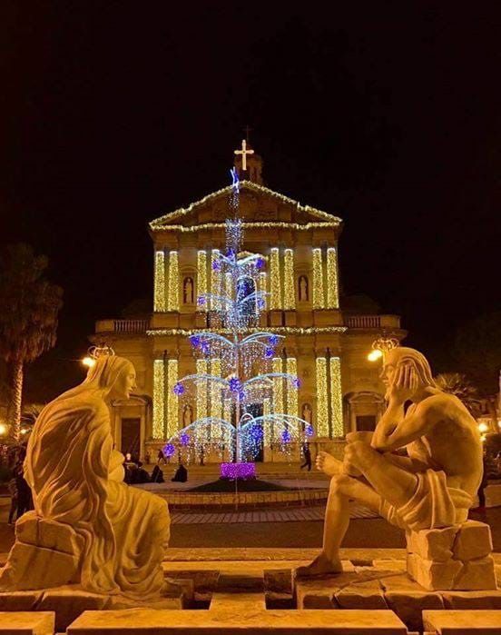 Chiesa illuminata di notte con luci blu e dorate, fiancheggiata da due statue di angeli in pietra sulla facciata.