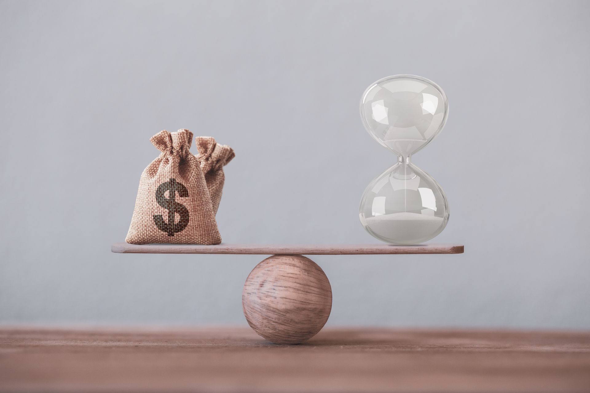 A bag of money and an hourglass are balancing on a wooden balance beam.