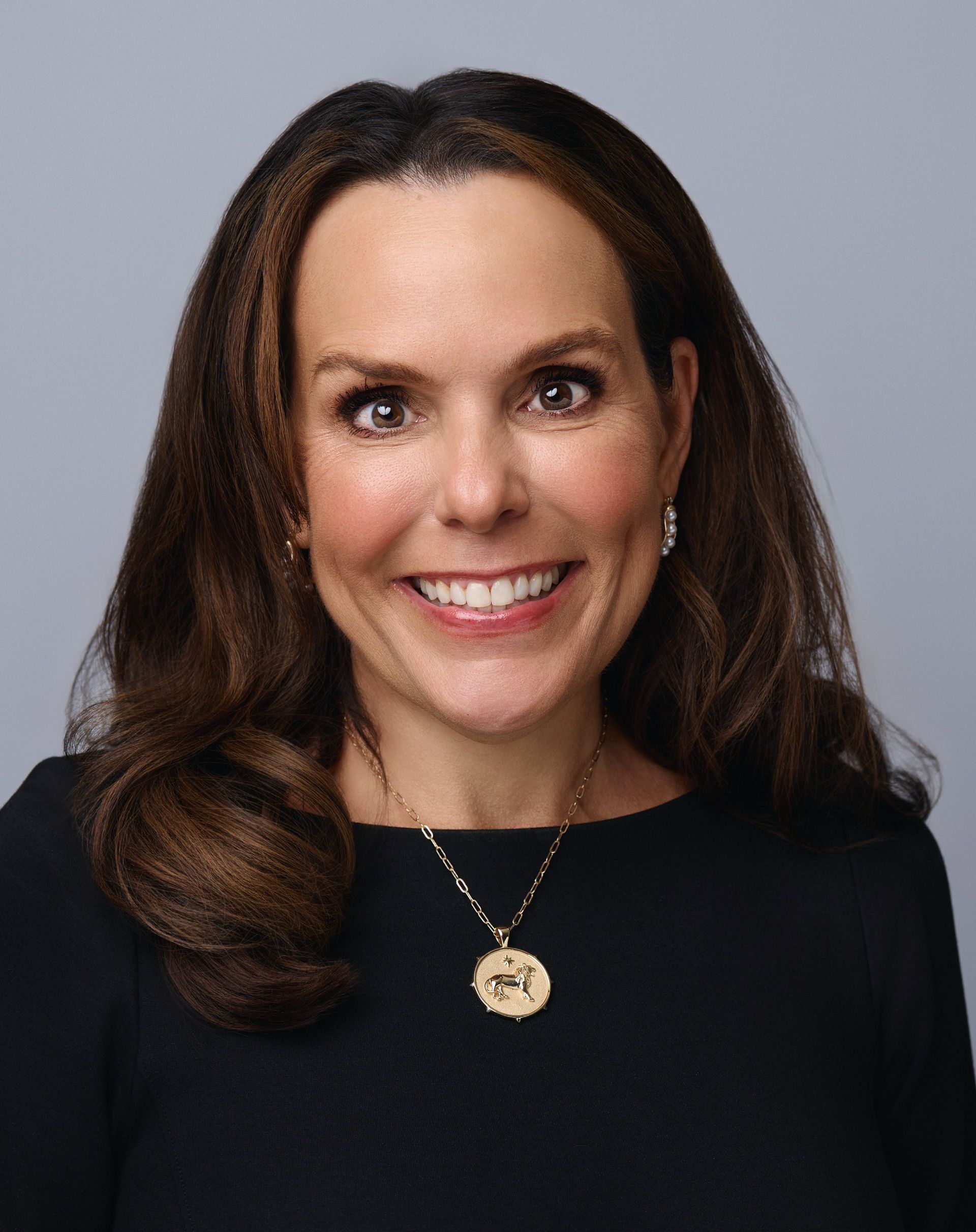 Woman with long brown hair, wearing a black dress and gold pendant necklace, smiling at the camera.