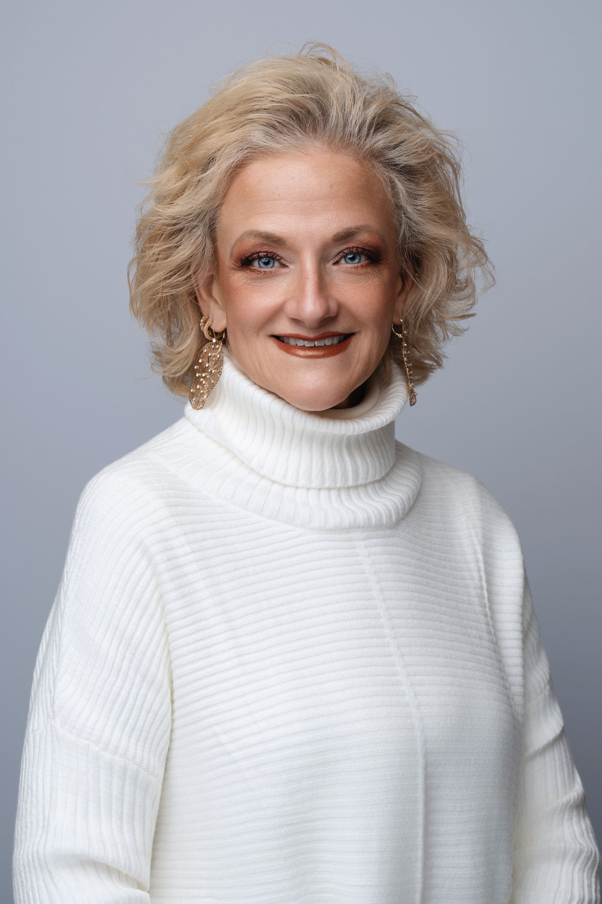 Woman with blonde curly hair, wearing a white turtleneck sweater, smiling at the camera.