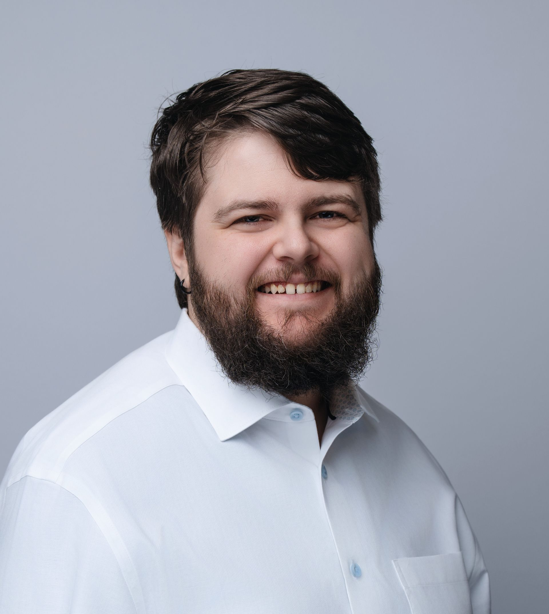 Man with dark hair and beard smiles, wearing a light blue button-up shirt against a grey background.