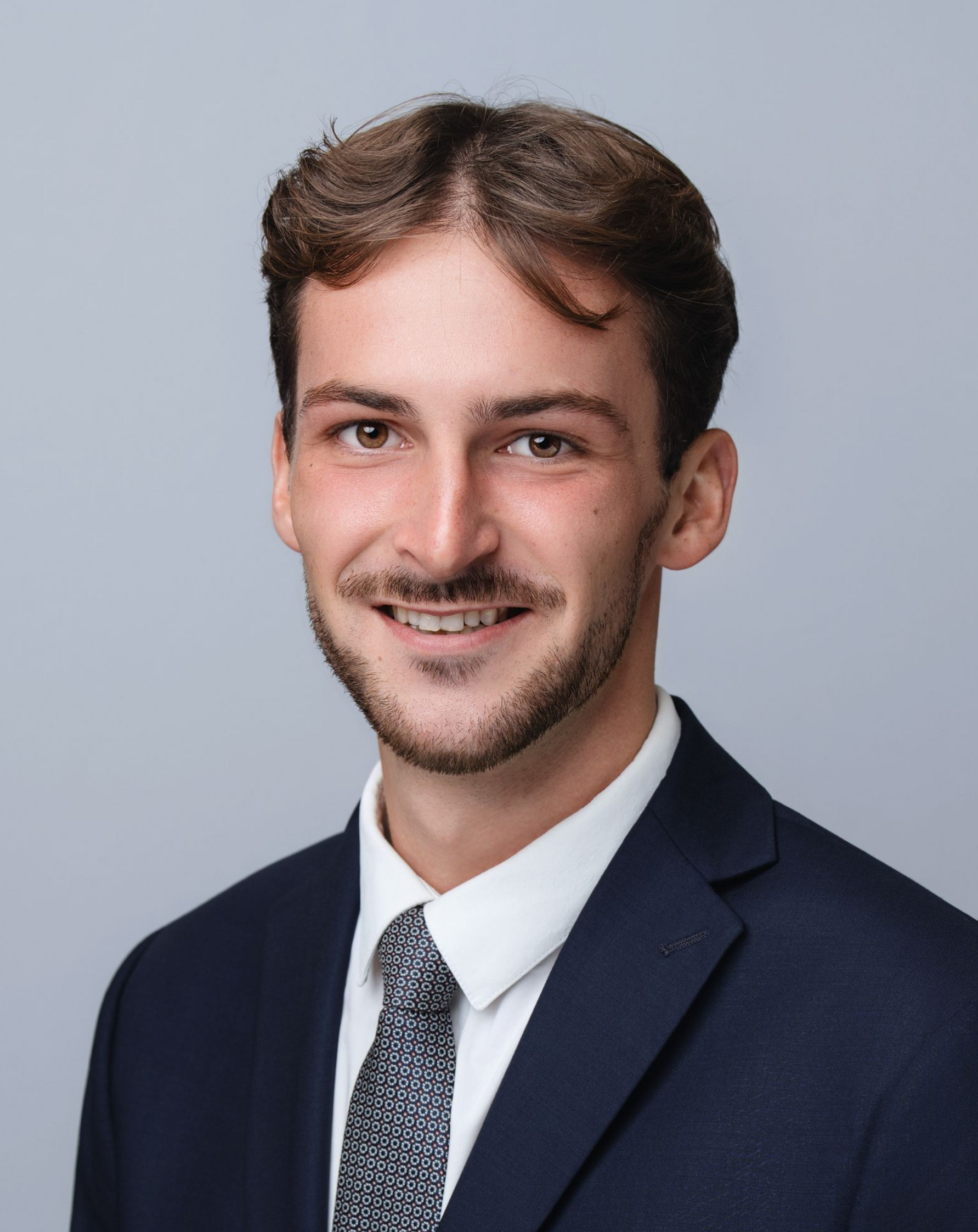 Man in dark suit, white shirt, patterned tie, smiling against a gray background.