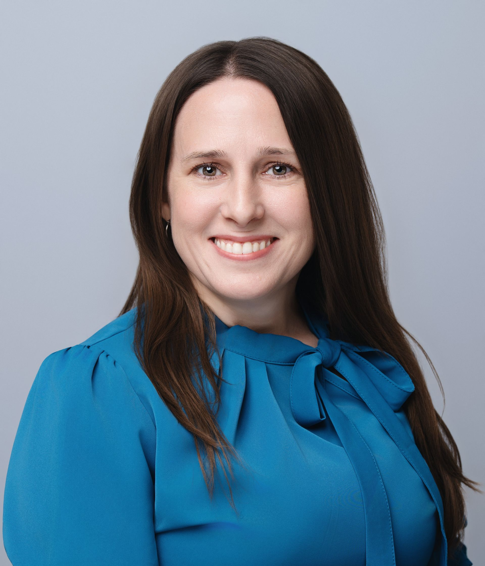 Woman with long brown hair, wearing a blue blouse, smiling against a gray background.