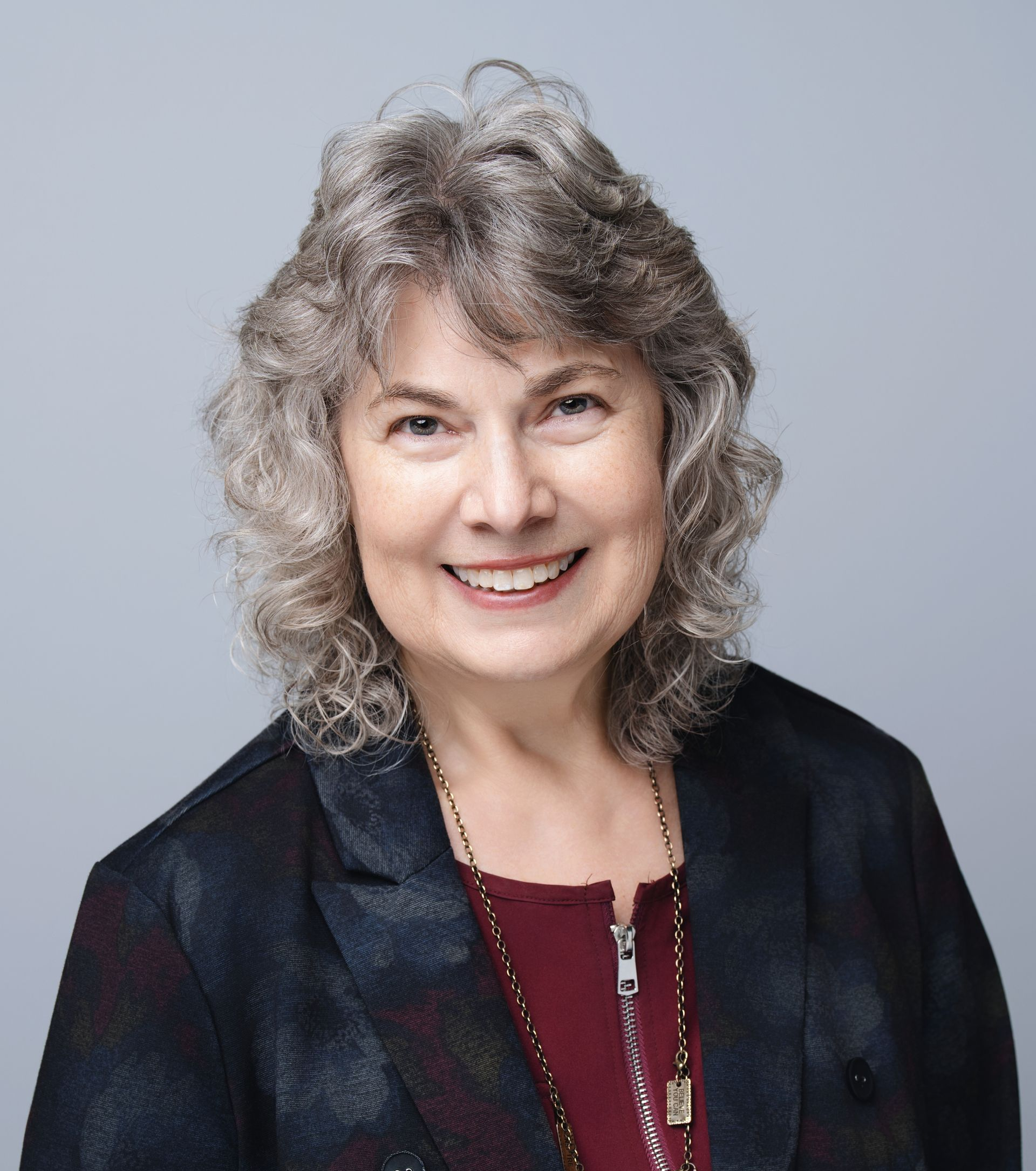 Woman with curly gray hair, smiling, wearing a dark blazer over a burgundy shirt, against a gray backdrop.