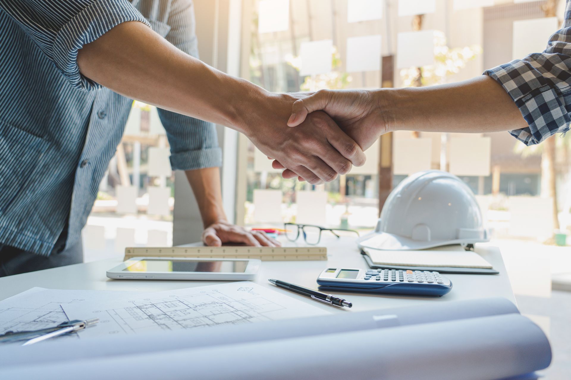 Handshake over blueprint on desk with hard hat and pen in a bright office